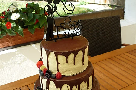 Vierstöckige Hochzeitstorte mit runterlaufender Schokoladenglasur und weißer Creme, verschiedenen Beeren auf drei Stockwerken und einem Brautpaar-Topper mit Schriftzug „Mr & Mrs“ oben drauf. Im Hintergrund ein Garten mit bepflanzten Blumentöpfen und grüner Wiese.