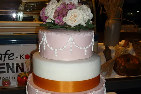 Vierstöckige Hochzeitstorte mit weißer und rosa Glasur, orangene-gelbem Band um zwei Stockwerke und einem rosa-weißen Blumenarrangement obenauf.