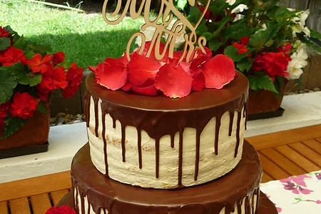 Dreistöckige Hochzeitstorte mit runterlaufender Schokoladenglasur auf allen drei Stockwerken, drei roten Rosen unten und dem Schriftzug „Mrs & Mrs“ aus Holz obenauf. Im Hintergrund ein Garten mit bepflanzten Blumentöpfen und grüner Wiese.