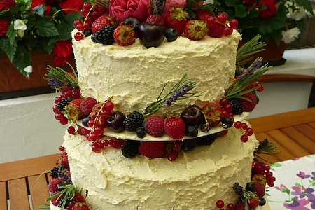Dreistöckige Torte mit weißer Creme, dekoriert mit Beeren, Kirschen, Lavendel und Rosmarinzweigen steht auf einem Tisch. Im Hintergrund ein Garten mit bepflanzten Blumentöpfen und grüner Wiese.