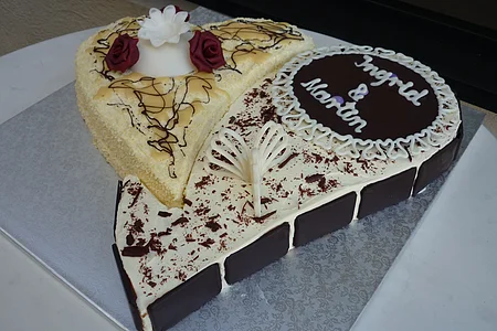Herzförmige Hochzeitstorte mit weißer und dunkler Schokolade, beschriftet mit „Ingrid & Martin“ und dekoriert mit zwei roten Rosen und verschiedener Schokoladen-Dekoration, steht auf einer silbernen Unterlage auf einem Tisch.