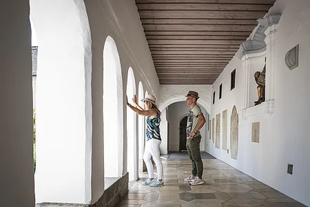 Zwei Personen mit Hüten stehen in einem Arkadengang, dem Kreuzgang im ehemaligen Kloster in Monheim mit Holzbalkendecke, weißen Wänden und Bogenfenstern, eine schaut durch die Bögen nach links hinaus.