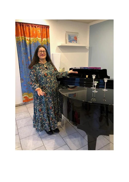 Frau mit langem Kleid und Brille steht neben schwarzem Flügelklavier in hellem Raum mit buntem Vorhang. Meditative Klänge am 18.01.26 in Beas Lädele in Monheim