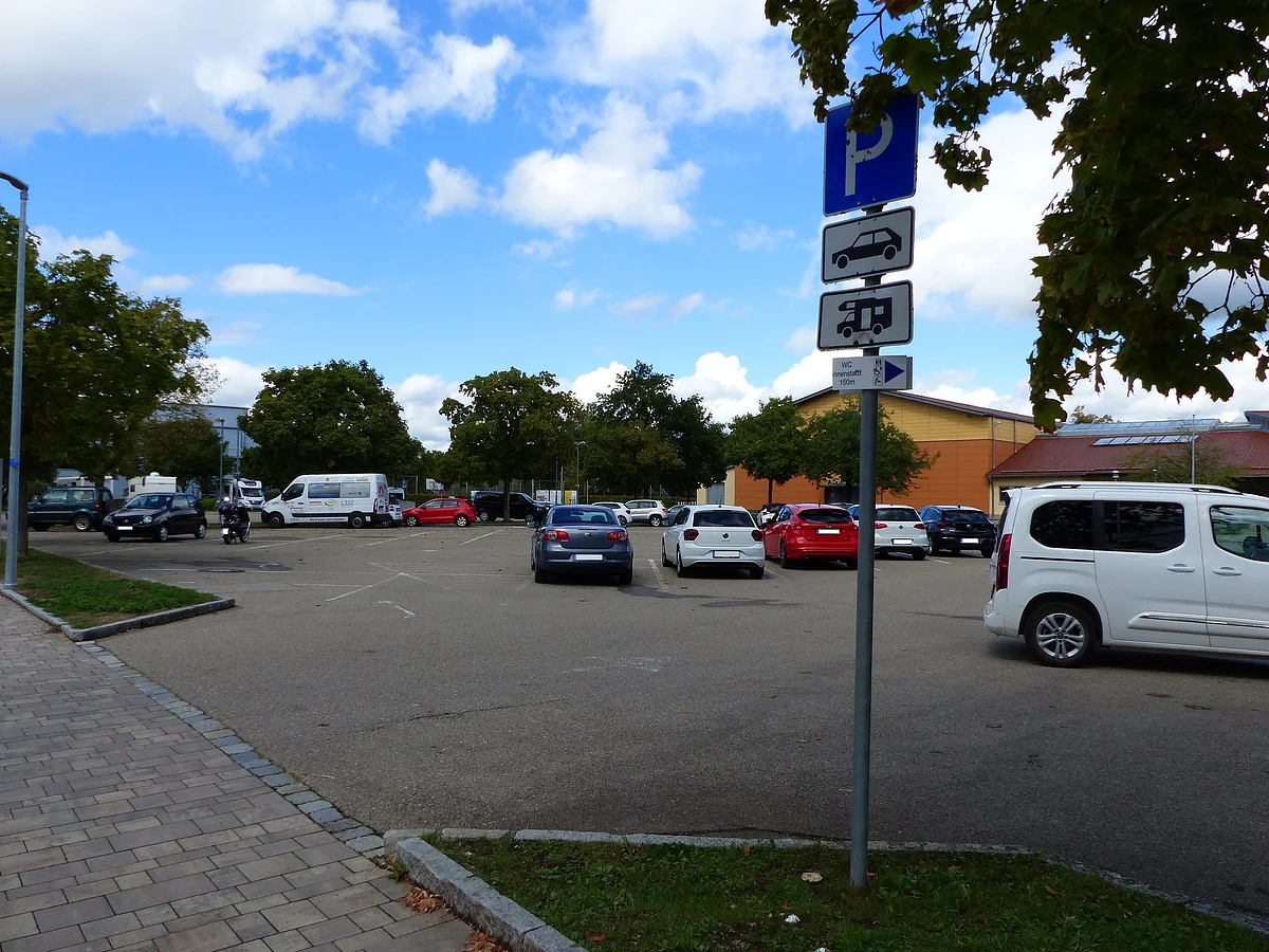 Parkplatz mit mehreren geparkten Autos und einem Schild für Pkw und Wohnmobile unter blauem Himmel mit Wolken. Im Hintergrund ein oranges Gebäude mit rotem Dach. Links im Bild ein Stück eines Gehweges.