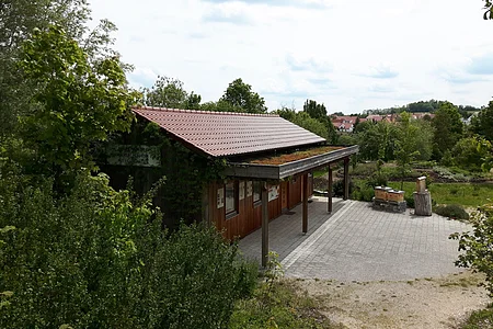Holzgebäude mit rotem Dach und begrüntem Vordach, umgeben von Bäumen und einem gepflasterten Vorplatz, bei bewölktem Himmel.