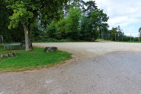 Parkplatz mit Kiesfläche, umgeben von Bäumen, links im Bild unter dem Baum eine Sitzgruppe aus Metall. Im Hintergrund eine Absperrung zum Fußballfeld unter bewölktem Himmel.