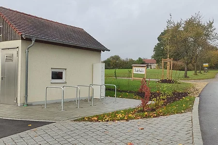 Kleines Gebäude mit Metalltür und kleinem Fenster, sowie fünf Fahrradständern auf gepflastertem Platz neben einer Straße und einem Schild mit Ortsnamen Flotzheim, unter bewölktem Himmel.