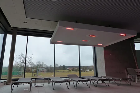 Innenraum eines Hallenbades mit Liegestühlen vor großen Fenstern und einer Deckenleuchte mit roten Lichtern.