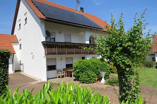 Zweistöckiges weißes Haus mit Solarpaneelen auf rotem Dach und grünem Garten bei klarem Himmel.