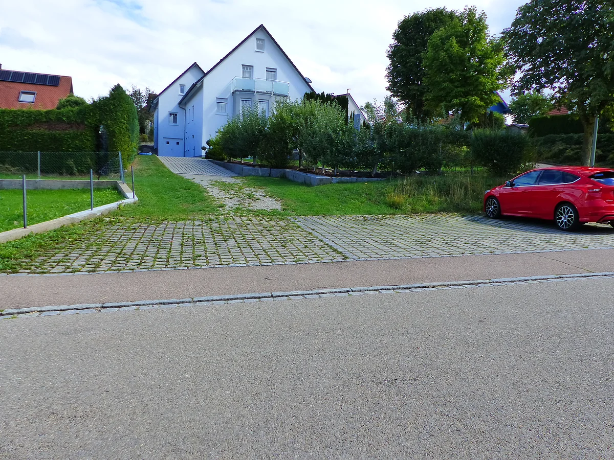 Zwei weiße Häuser mit Giebeldach im Hintergrund, davor Rasenflächen, Bäume und ein rotes Auto auf gepflastertem Parkplatz.