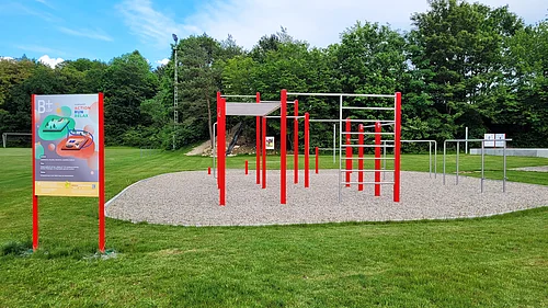 Outdoor-Fitnessbereich mit roten Reckstangen und Klettergerüst auf Kiesfläche, umgeben von Rasen und Bäumen