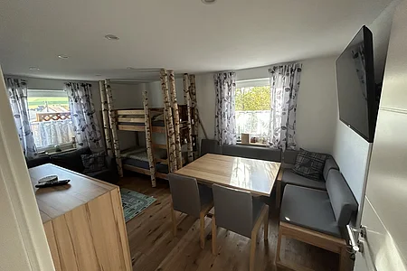 Wohnzimmer mit Holzboden, Eckbank, Esstisch, Etagenbett aus Holzstämmen und zwei Fenstern mit gemusterten Vorhängen.