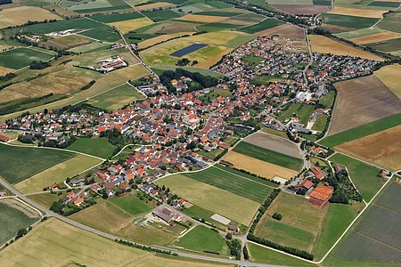 Luftaufnahme eines Dorfes mit roten Dächern, umgeben von landwirtschaftlichen Feldern und Straßen.
