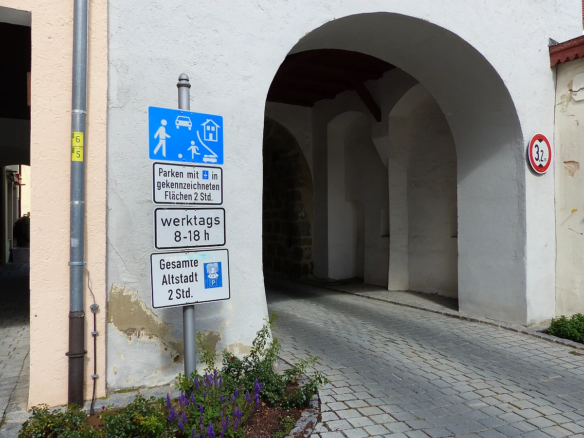Verkehrszeichen mit Parkhinweisen und Fußgängerzone vor einem weißen Torbogen in einer gepflasterten Straße. Vor dem Torbogen ein kleines bepflanztes Blumenbeet.