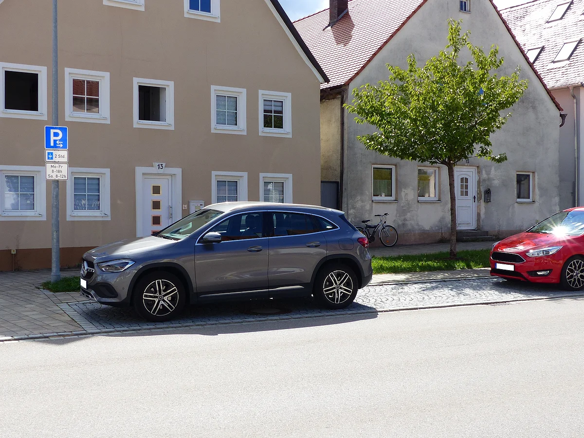 Zwei parkende Autos stehen vor Wohnhäusern auf einem gepflasterten Parkplatz mit dem blauem Parkplatzschild und darunter ein Schild mit zeitlicher Begrenzung. Ein Baum steht zwischen den beiden Autos auf einer Grünfläche. Im Vordergrund eine Straße.