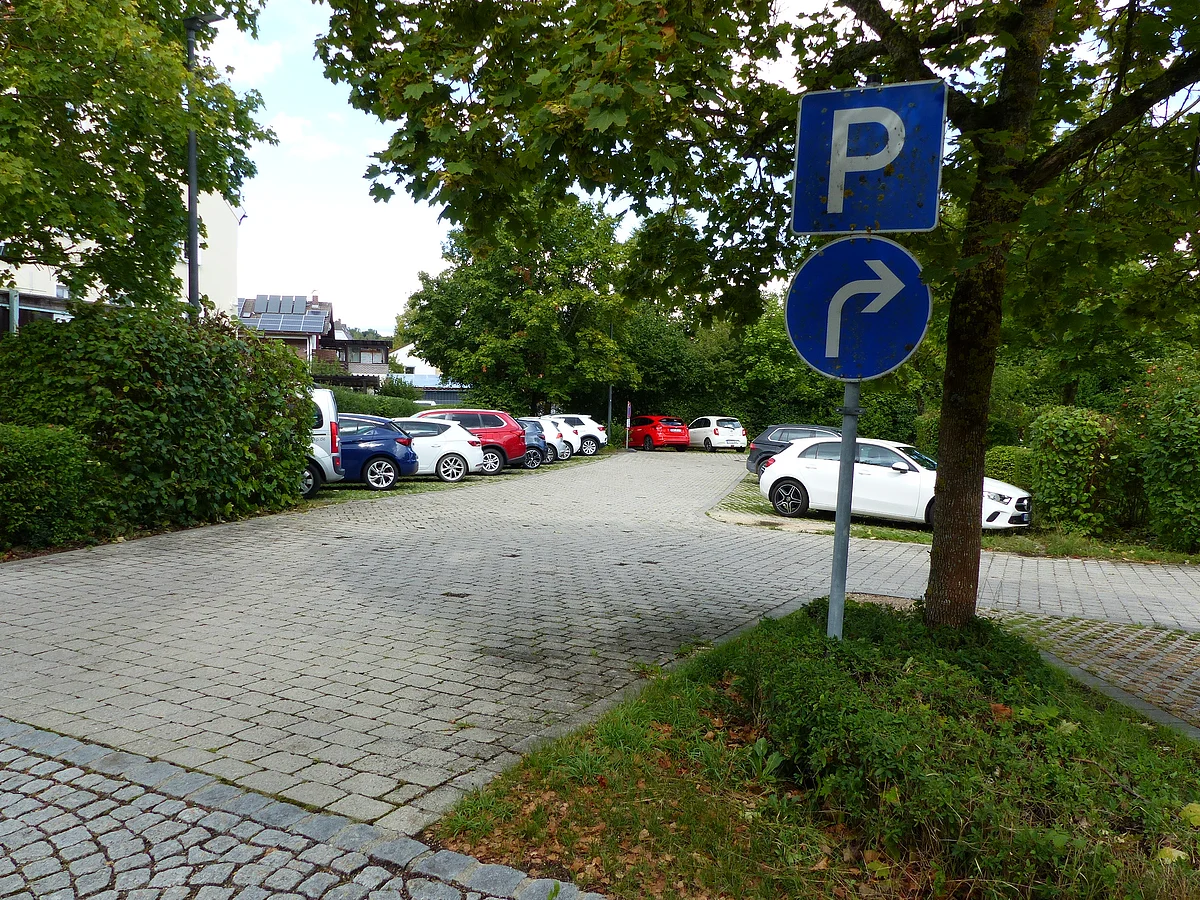 Parkplatz mit mehreren geparkten Autos und zwei Verkehrsschildern für Parken und Rechtsabbiegen unter Bäumen bei Tageslicht.