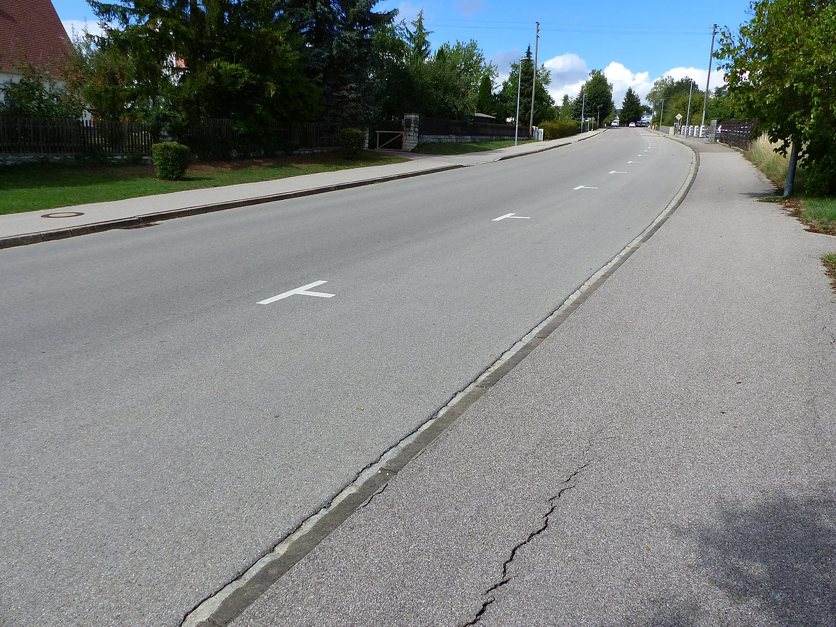 Leere Straße mit einem Gehweg links im Bild und weißen Parkmarkierungen auf der rechten Seite unter blauem Himmel und Bäumen am Straßenrand.