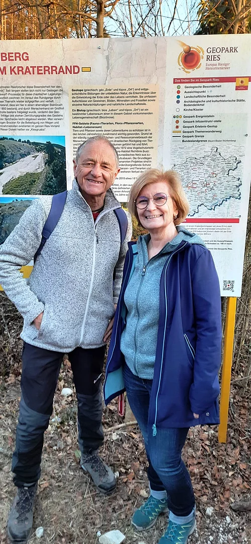 Zwei ältere Personen in Outdoorbekleidung stehen vor einem Informationstafel zum Geopark Ries im Wald.