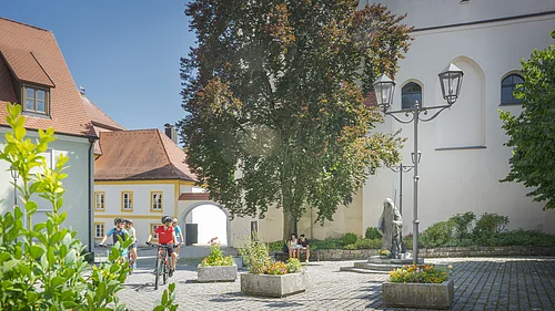 Radfahrer unterwegs durch die Historische Innenstadt von Monheim Innenhof mit Radfahrern, Baum, Sitzbank, Statue und historischen Gebäuden bei Sonnenschein