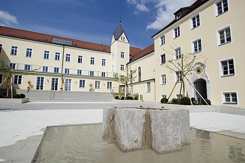 Stiftung Sankt Johannes in Schweinspoint - ehemaliges Schloss Innenhof mit modernem Brunnen und gelben Gebäuden mit roten Dächern unter blauem Himmel
