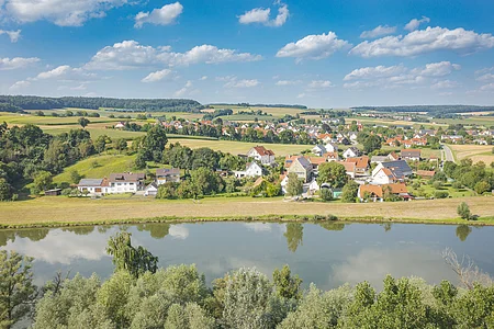 Dorf mit Häusern, Feldern und Bäumen an einem Fluss unter blauem Himmel mit Wolken