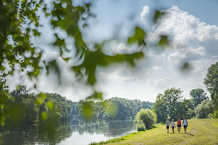 Vier Personen gehen auf einem Grasweg entlang eines Flusses unter blauem Himmel mit Wolken spazieren. Unscharfer Ast eines Baumes im Vordergrund.