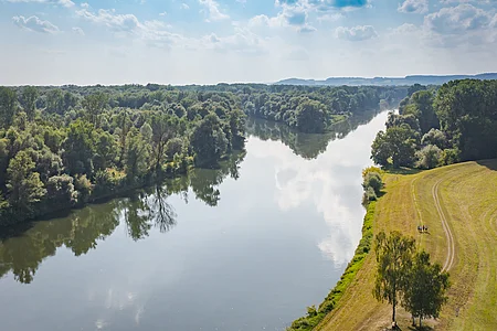 Fluss mit bewaldetem Ufer links und Wiese mit Wanderern rechts unter bewölktem Himmel