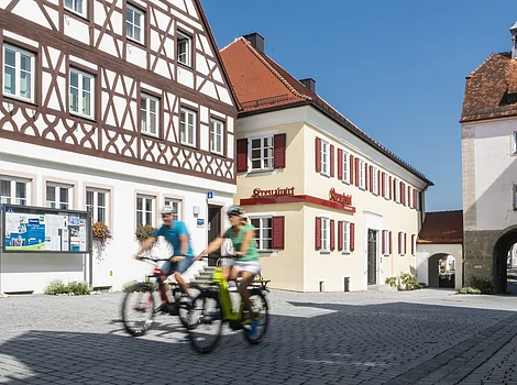 Fachwerkhaus und historische Gebäude an sonnigem Platz mit zwei Radfahrern in Bewegung