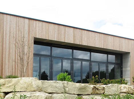 B+ Natur- und Umweltbildungszentrum Blossenau Flaches Holzgebäude mit großen Fenstern, davor Steinmauer und bunte Wildblumenwiese mit roten Mohnblumen.
