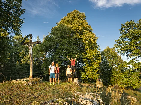 Familie mit zwei Kindern steht auf einer Wiese neben einem Holzkreuz mit Jesusfigur bei Sonnenschein.