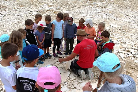 Kinder stehen im Kreis um eine Person mit rotem Shirt und Hut, die auf dem Boden sitzend Steine bearbeitet.