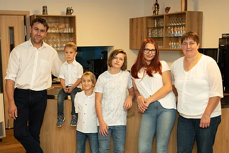 Sechs Personen, drei Erwachsene und drei Kinder, stehen und sitzen in einem Raum mit Holzboden und Holztheke.