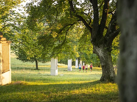 Vier Personen gehen auf einer Wiese mit Bäumen und mehreren weißen Säulen entlang.