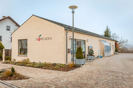 Eingang eines hellen Gebäudes mit Schriftzug „Dorfladen“ und Pflanzkübeln vor der Tür.