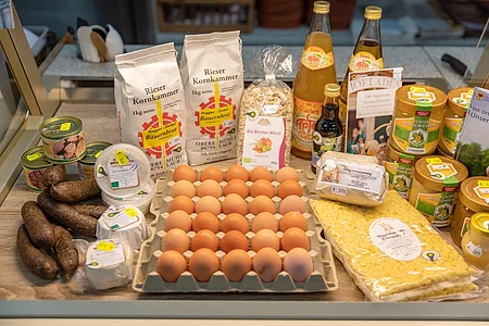 Verschiedene Lebensmittel wie Eier, Honig, Wurst, Brot, Müsli und Getränke auf einem Holztisch.