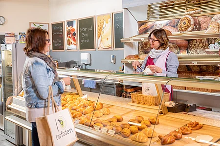 Kundin mit Tasche vor Theke mit Backwaren spricht mit Verkäuferin in Bäckerei mit Regalen voller Brot.