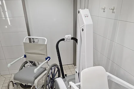 Weißer Duschrollstuhl und weißer Badelift für einen Pool mit blauer Sicherung im gefliesten Raum eines Hallenbads vor geschlossener Tür.