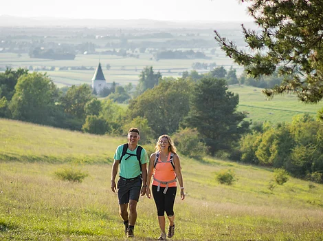 Zwei Wanderer mit Rucksäcken gehen auf einem Weg durch eine grüne Wiese mit Bäumen und Dorf im Hintergrund.