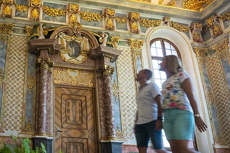 Zwei Personen gehen in einem prunkvoll verzierten Raum mit goldenen Stuckelementen und bemalter Decke.