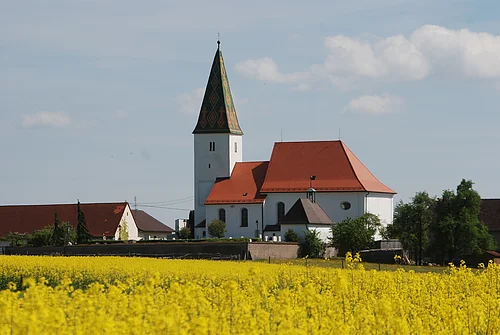 Kirche mit rotem Dach und Turm hinter einem gelben Rapsfeld unter blauem Himmel mit Wolken