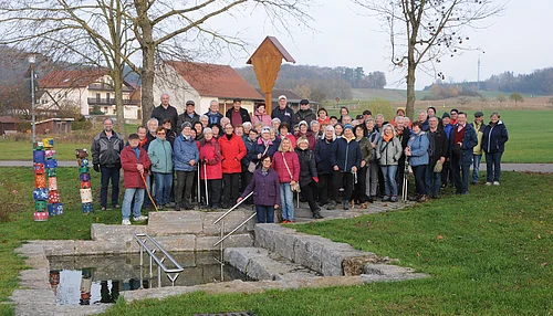 Große Gruppe älterer Menschen steht im Park vor einem kleinen Wasserbecken mit Treppe und Geländer. Monatliche Wanderung für Senioren vom Verein Soziales Miteinander