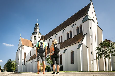 Zwei Personen mit Rucksäcken stehen vor einer großen weißen Kirche mit Turm und blauem Himmel.