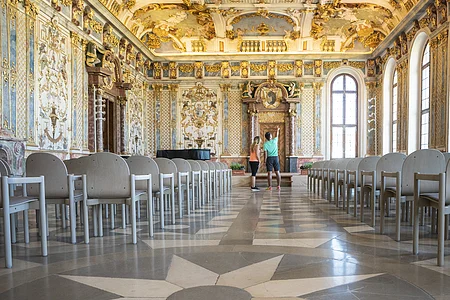 Prächtig verzierter Saal mit goldenen Stuckverzierungen, Sternmuster auf dem Boden und zwei Personen in der Mitte.