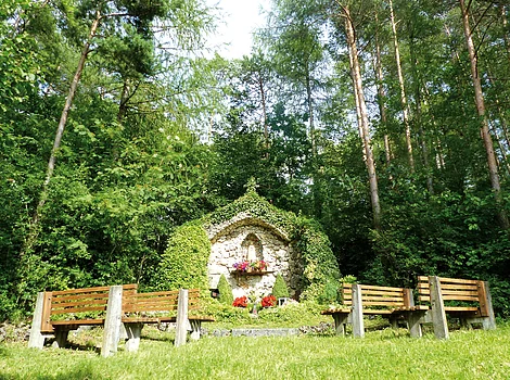 Steinernes Marienheiligtum mit Blumen, umgeben von Büschen und vier Holzbänken auf Grasfläche im Wald.