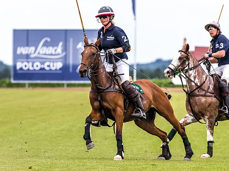Zwei Polospieler auf Pferden in Aktion auf einem Spielfeld mit Werbebanner im Hintergrund.