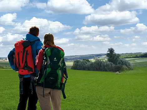 Zwei Personen mit Rucksäcken stehen auf einer grünen Wiese und blicken auf eine Landschaft mit blauem Himmel und Wolken.