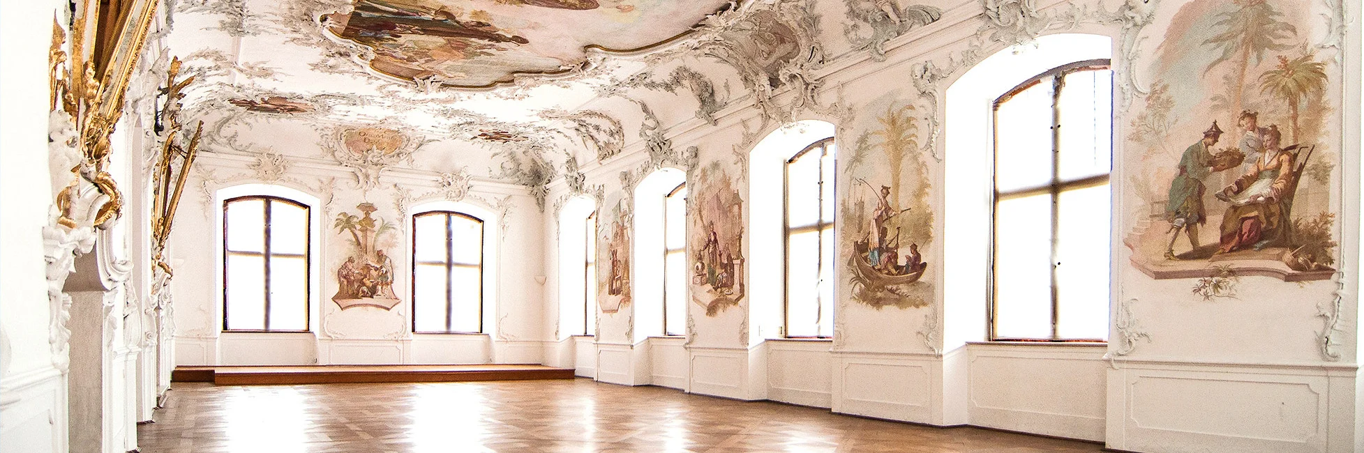 Großer heller Raum mit Stuckdecke, Deckengemälde und großen Fenstern an der rechten Wand.