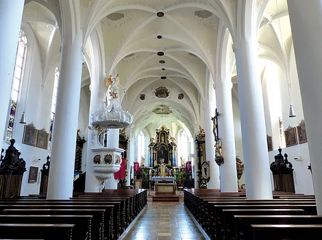 Innenraum einer Kirche mit weißen Säulen, Holzbänken und einem Altar am Ende des Mittelgangs