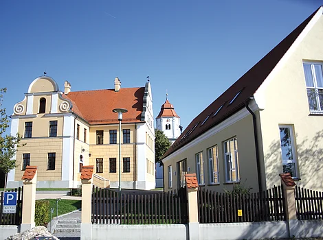 Gelbes historisches Gebäude mit rotem Dach und Kirchturm im Hintergrund bei klarem Himmel.