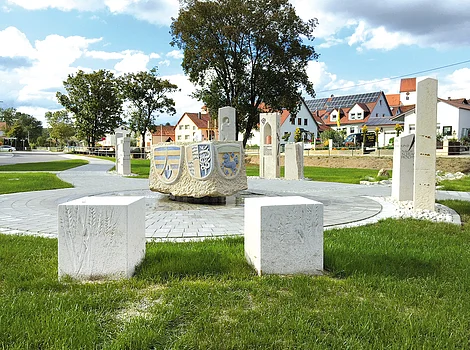 Steinskulpturen und Wappen in einem Kreis auf einer Wiese vor Häusern unter blauem Himmel mit Wolken.